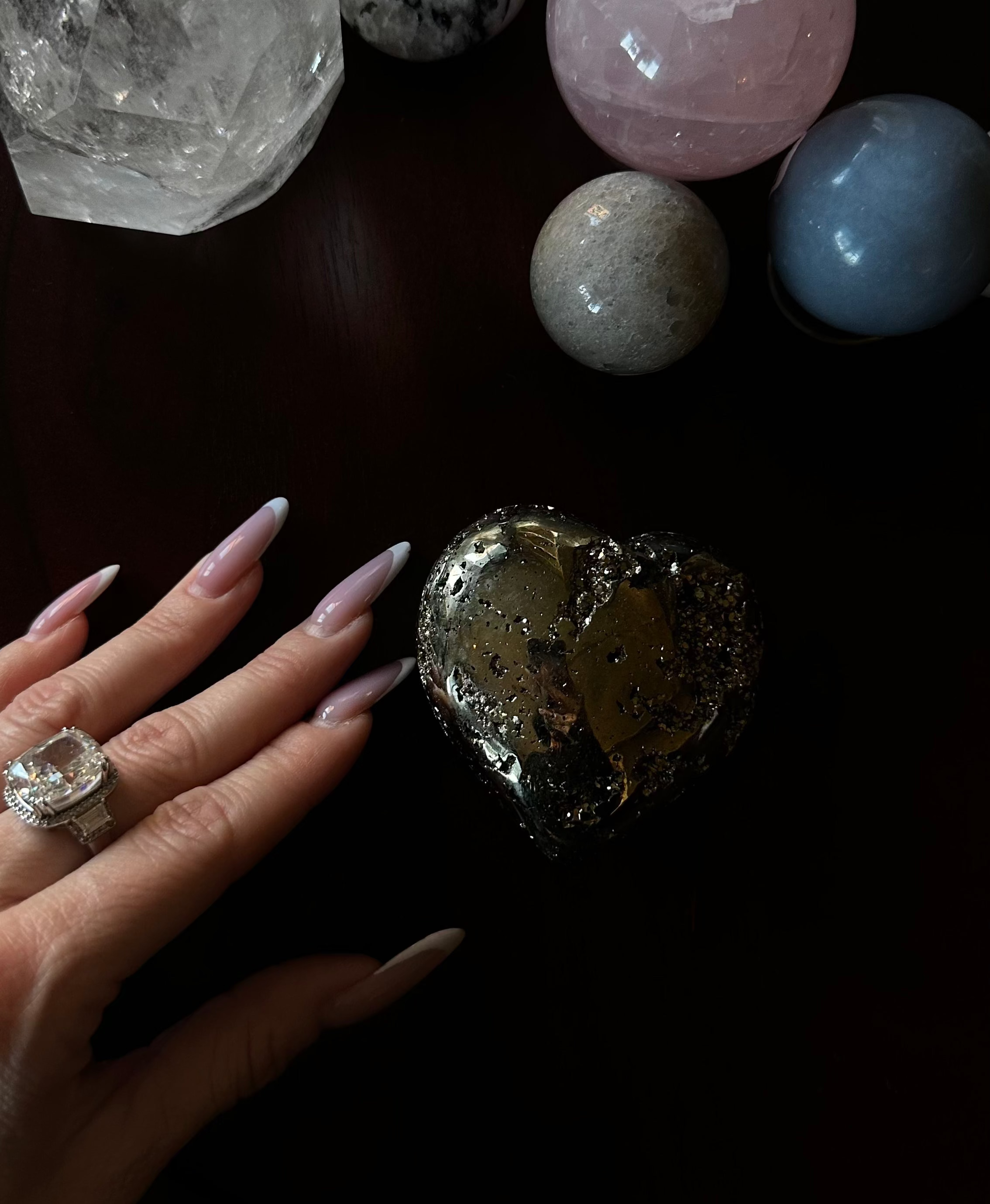 Heart-shaped pyrite crystal held by a hand with a ring, surrounded by various crystals on a dark surface